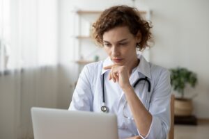 A female doctor in a white coat is looking at her laptop