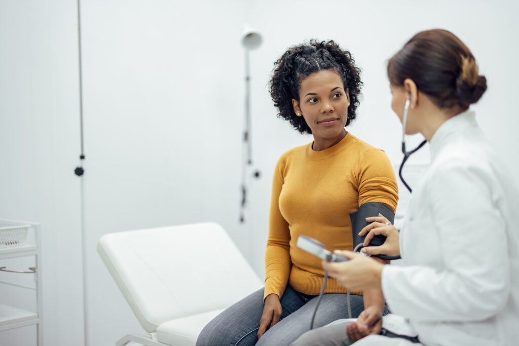 A female patient is sitting on an examination table, getting her blood pressure taken by a female doctor in a white lab coat.