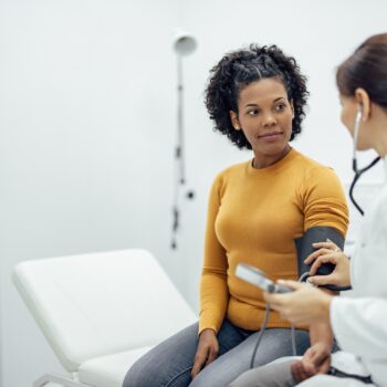 A female patient is sitting on an examination table, getting her blood pressure taken by a female doctor in a white lab coat.