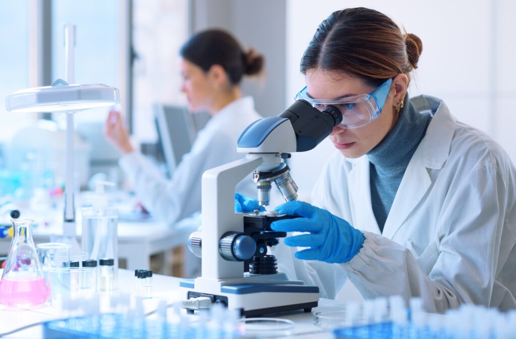 A female scientist in a white lab coat is looking at a sample through a microscope