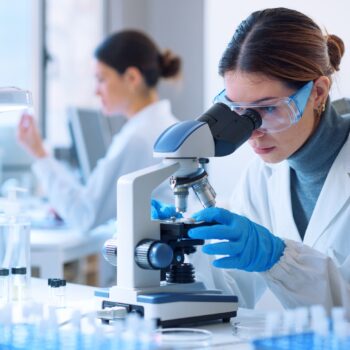 A female scientist in a white lab coat is looking at a sample through a microscope